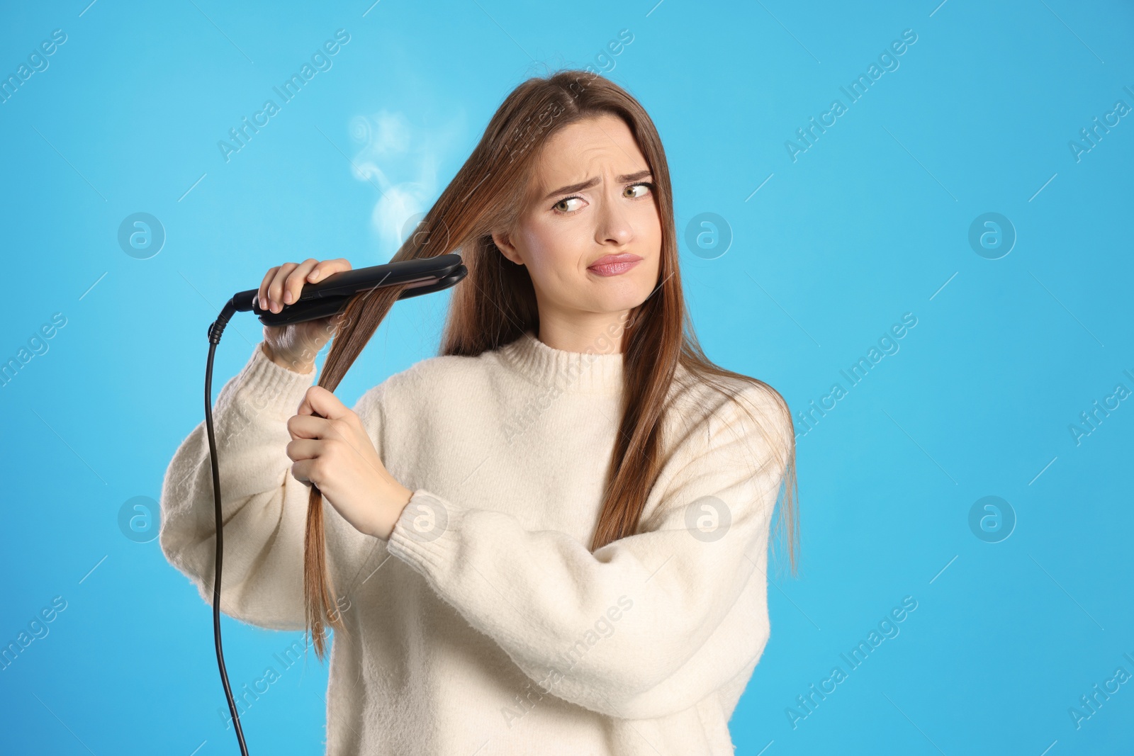 Image of Upset young woman with flattening iron on light blue background. Hair damage