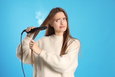 Image of Upset young woman with flattening iron on light blue background. Hair damage
