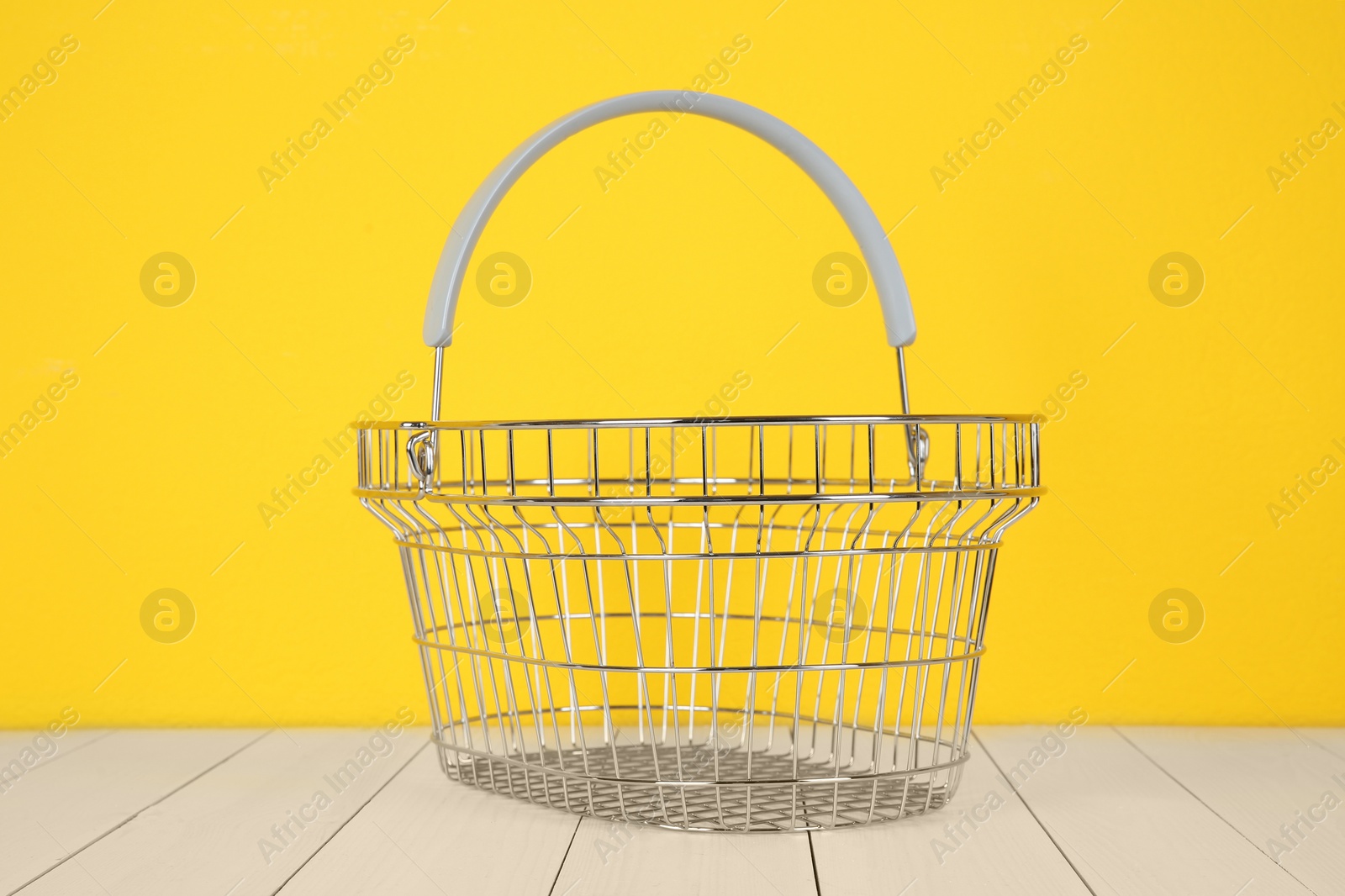 Photo of Empty metal shopping basket on white wooden table against yellow background