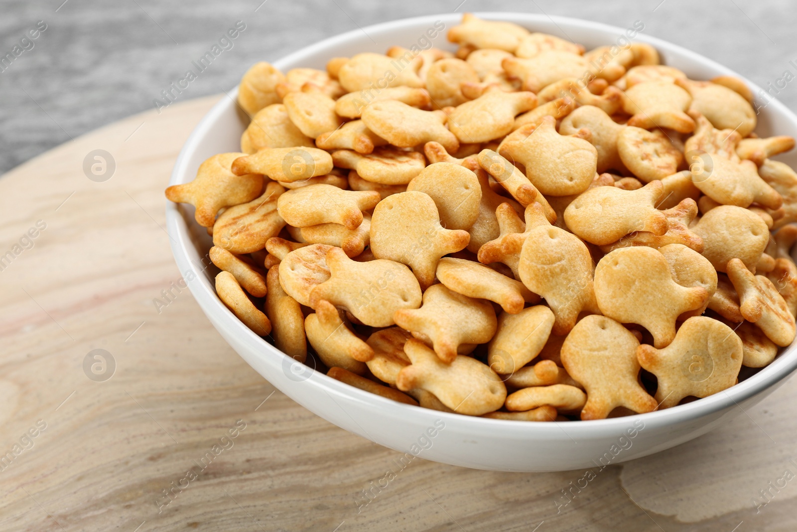 Photo of Delicious goldfish crackers in bowl on wooden board, closeup