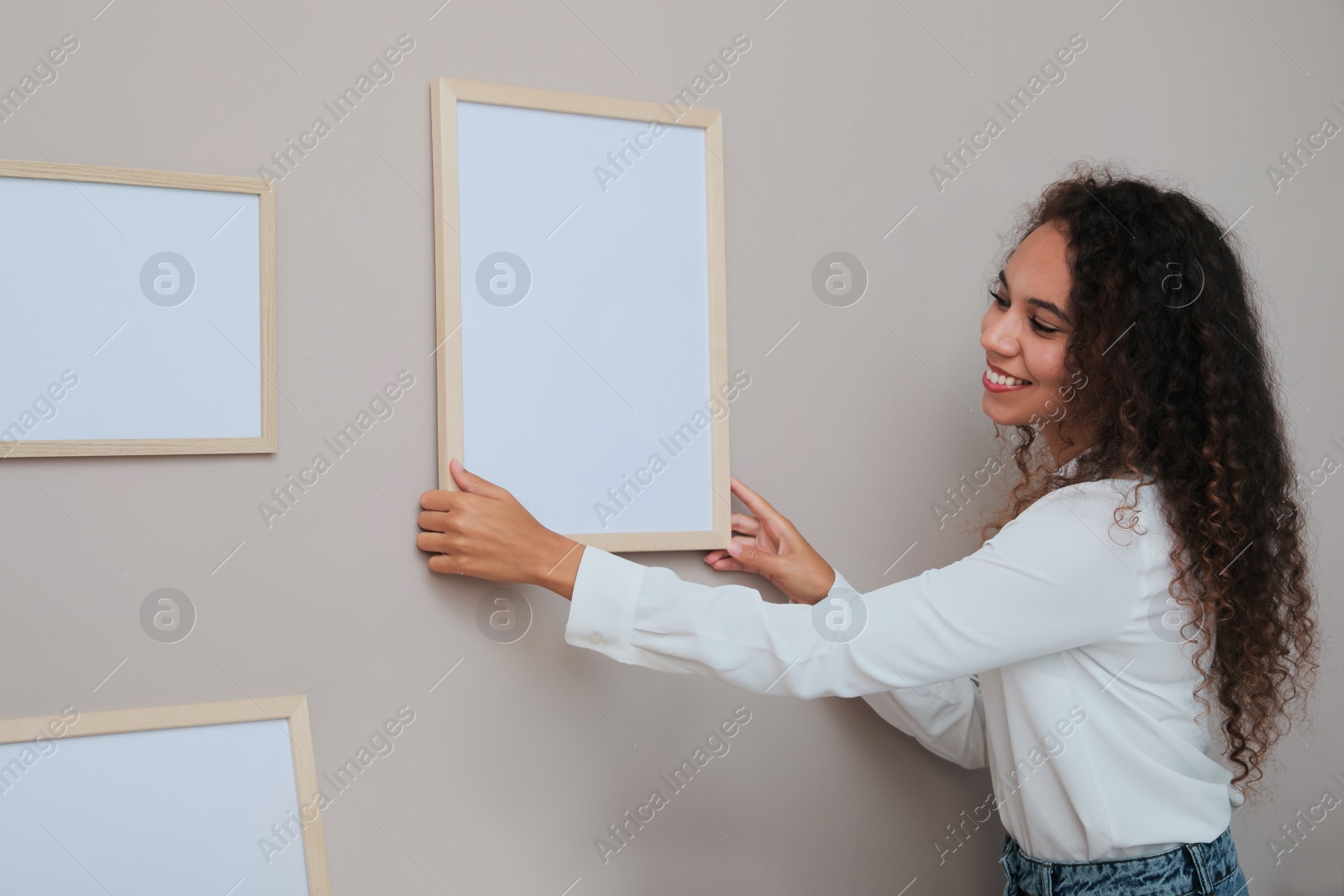 Photo of Happy African American woman hanging empty frame on pale rose wall in room. Mockup for design