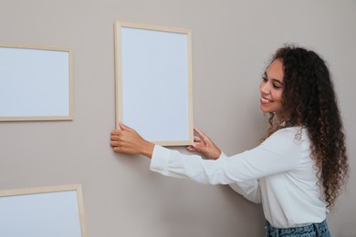 Happy African American woman hanging empty frame on pale rose wall in room. Mockup for design