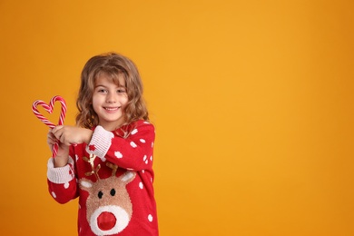 Photo of Cute little girl in Christmas sweater making heart shape with candy canes against orange background. Space for text