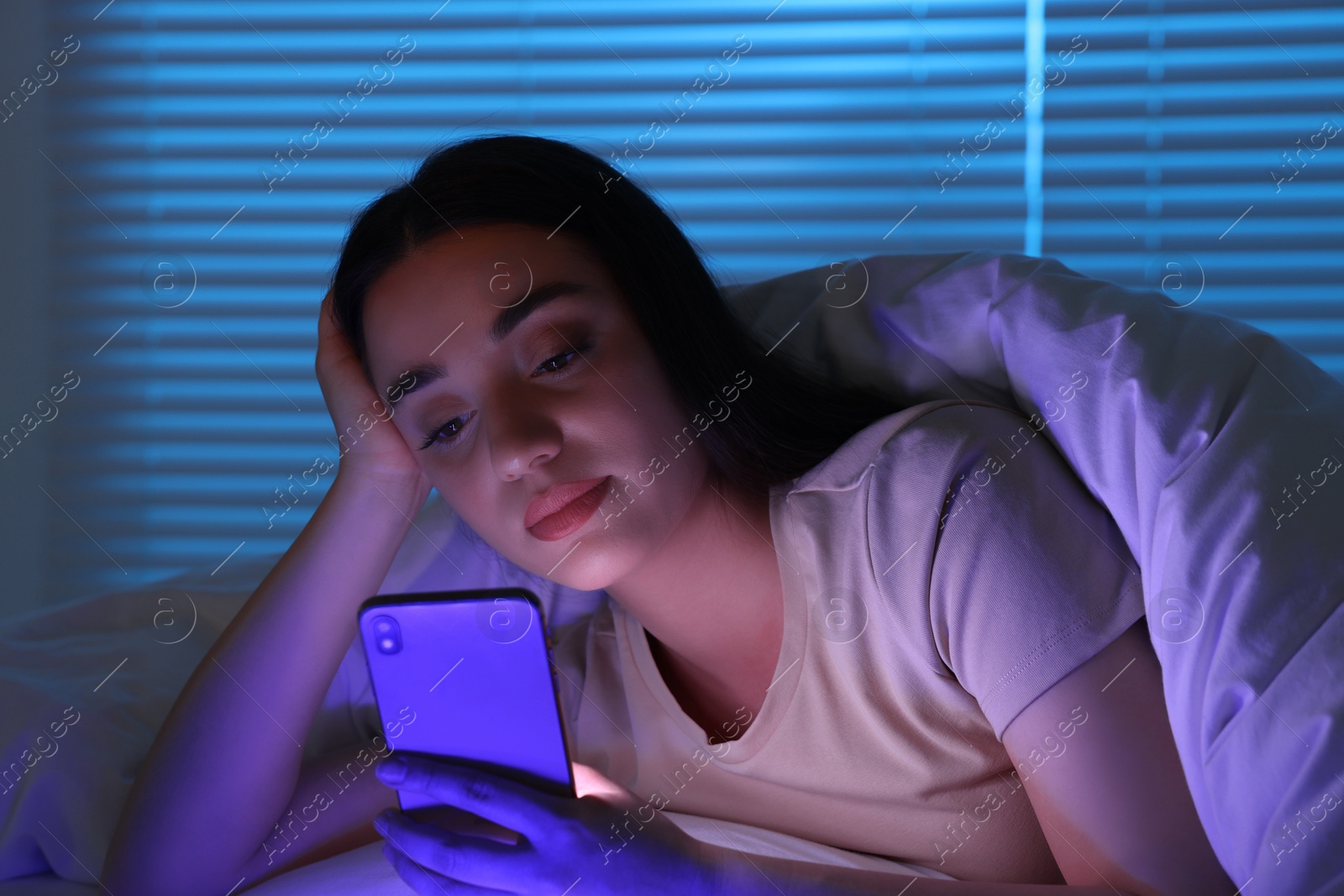 Photo of Young woman using modern smartphone in bed at night. Internet addiction