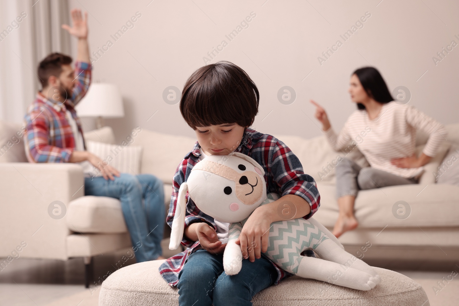 Photo of Sad little boy with toy and his arguing parents on sofa in living room