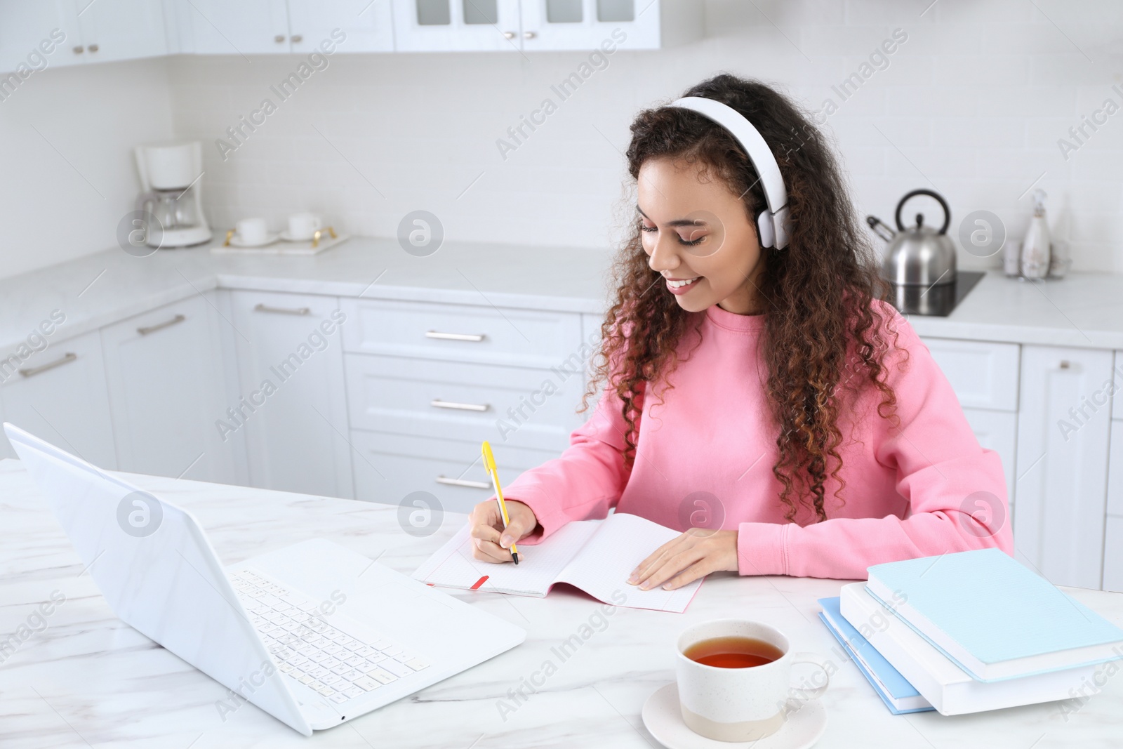 Photo of African American woman with modern laptop and headphones studying in kitchen. Distance learning