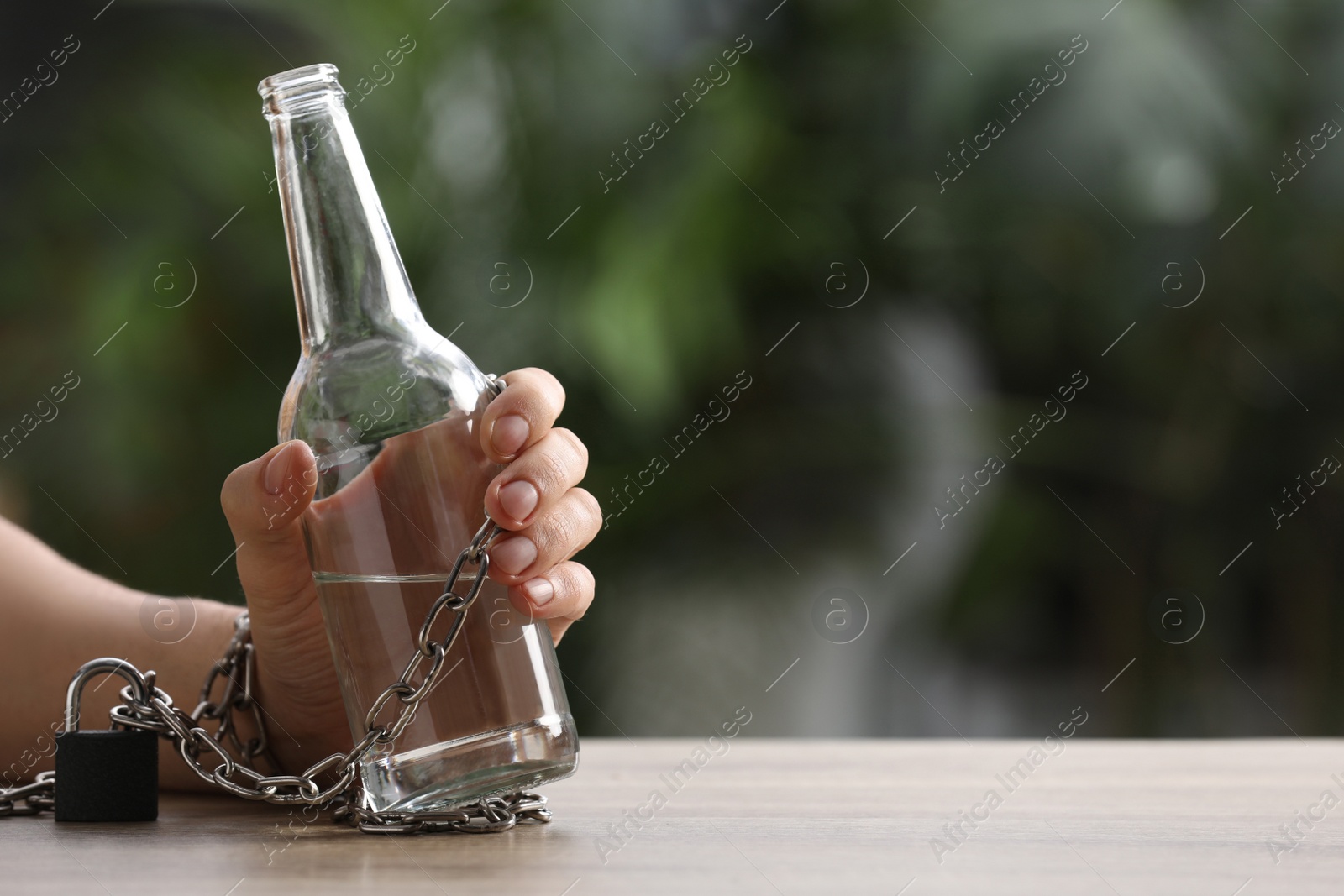 Photo of Man chained to bottle of vodka at table against blurred background, closeup with space for text. Alcohol addiction