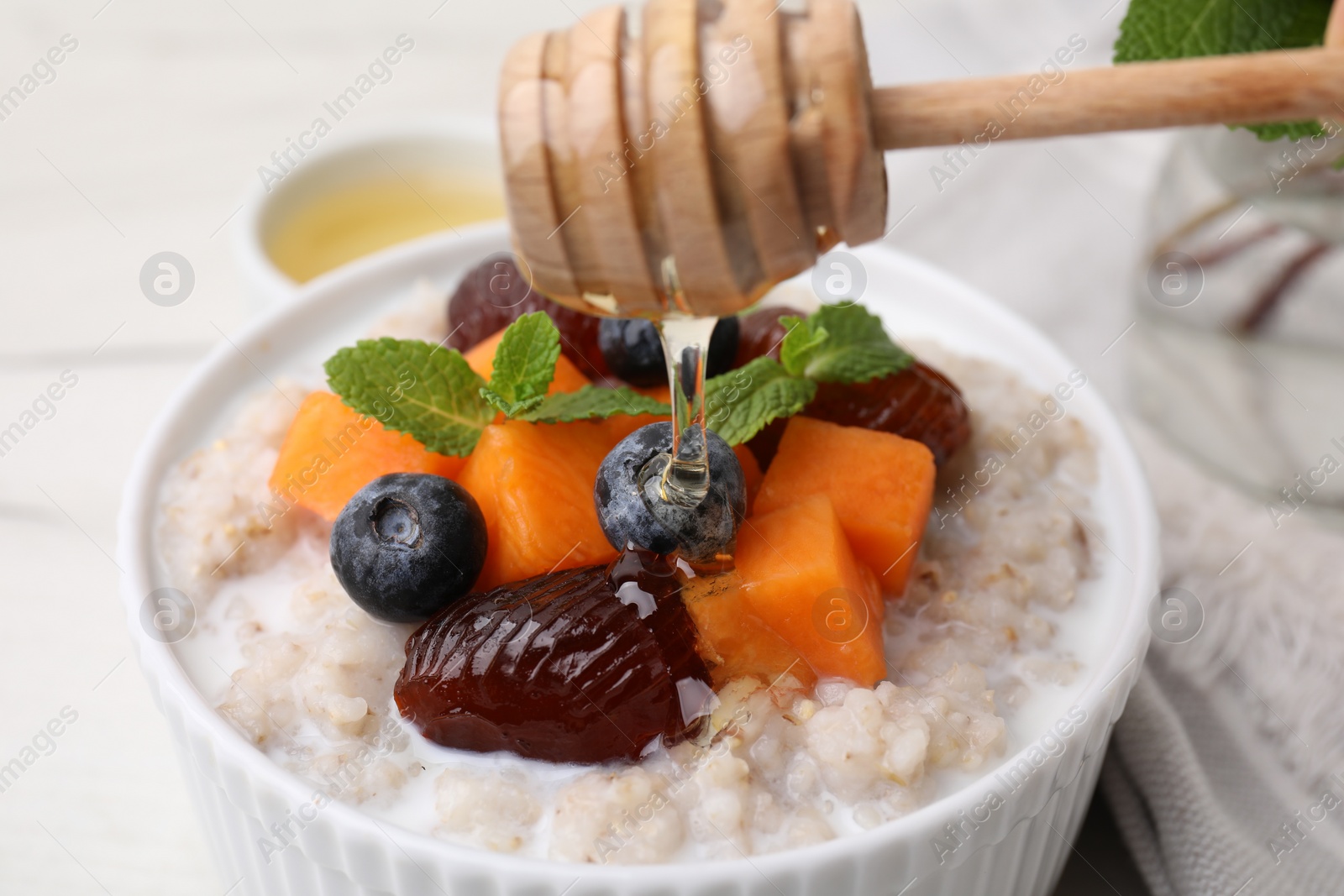 Photo of Pouring honey onto bowl of delicious barley porridge with blueberries, pumpkin, dates and mint at white table, closeup