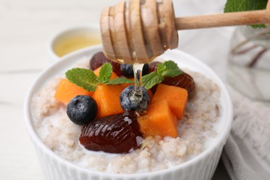 Pouring honey onto bowl of delicious barley porridge with blueberries, pumpkin, dates and mint at white table, closeup