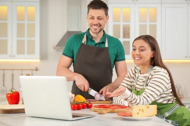 Happy couple making pizza together while watching online cooking course via laptop in kitchen