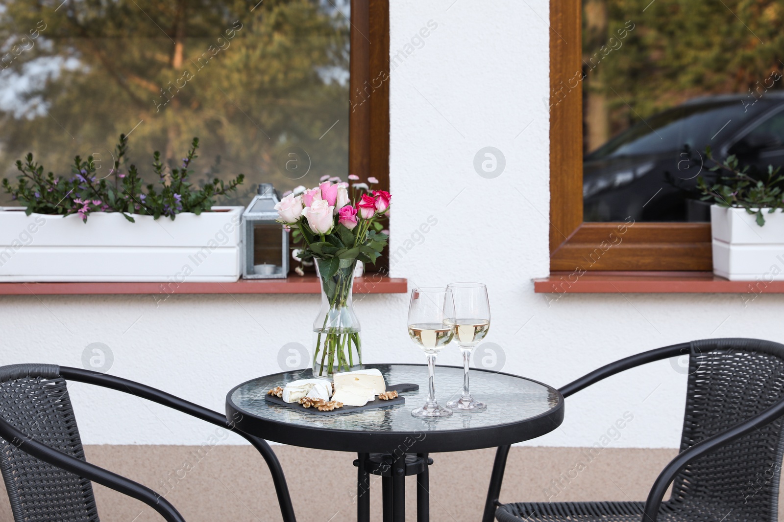 Photo of Vase with roses, glasses of wine and food on glass table near house on outdoor terrace