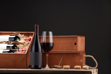 Box with wine bottles, glass, corks and corkscrew on wooden table against black background