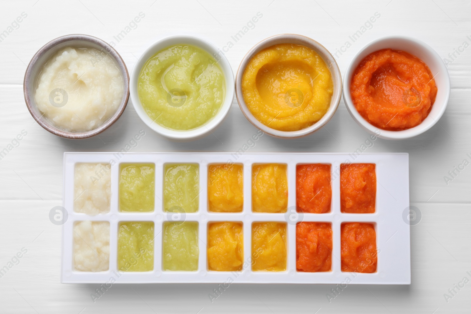 Photo of Different purees in ice cube tray and bowls on white wooden table, flat lay. Ready for freezing