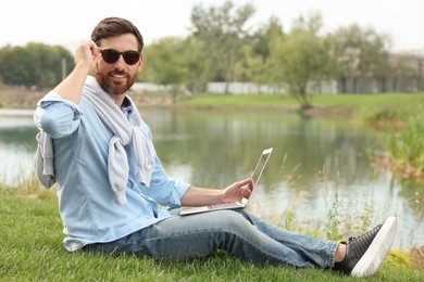 Man in sunglasses with laptop on green grass near lake