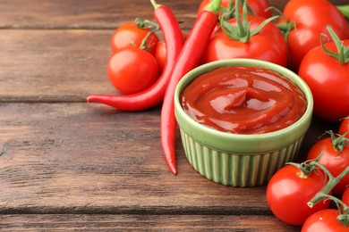 Bowl of tasty ketchup, chili peppers and tomatoes on wooden table, closeup. Space for text