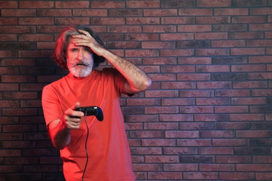 Emotional mature man playing video games with controller near brick wall. Space for text