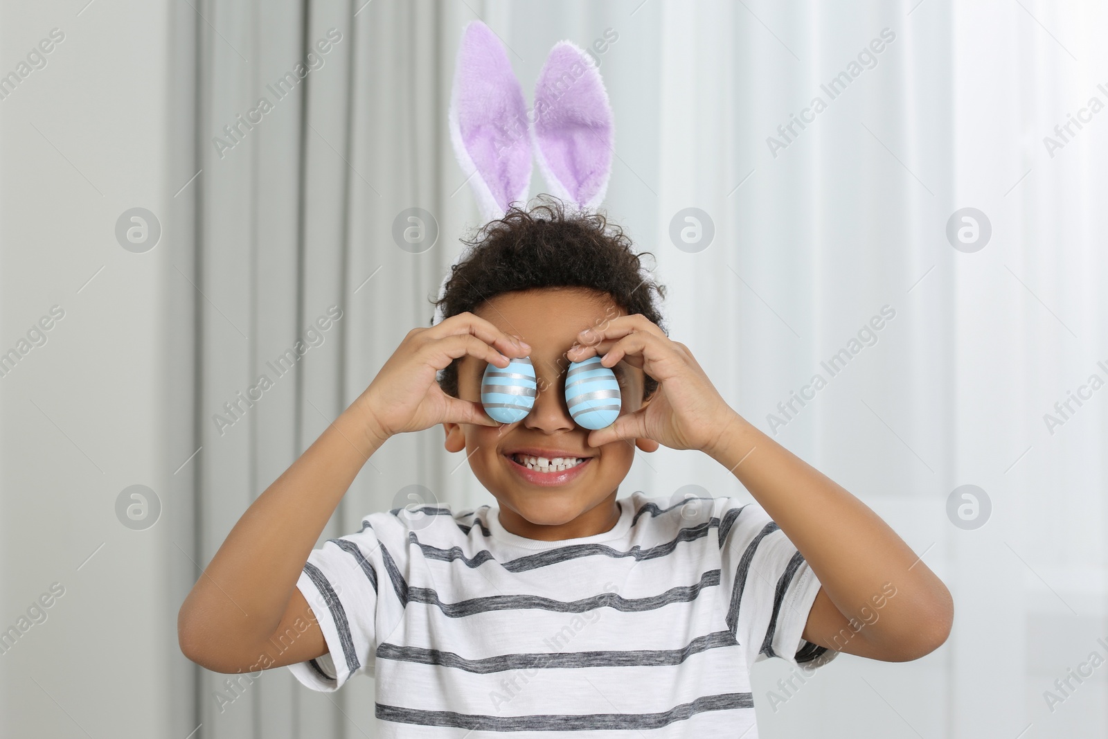 Photo of Cute African American boy in bunny ears headband covering eyes with Easter eggs indoors