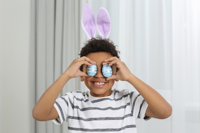 Photo of Cute African American boy in bunny ears headband covering eyes with Easter eggs indoors