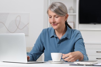 Beautiful senior woman with cup of drink using laptop at white table indoors
