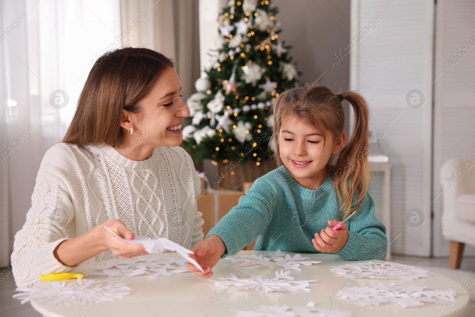 Photo of Happy mother and daughter making paper snowflake at table near Christmas tree indoors