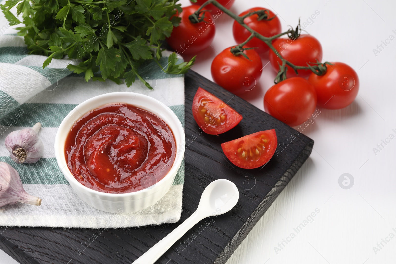 Photo of Organic ketchup in bowl, fresh tomatoes, parsley and garlic on white table. Tomato sauce