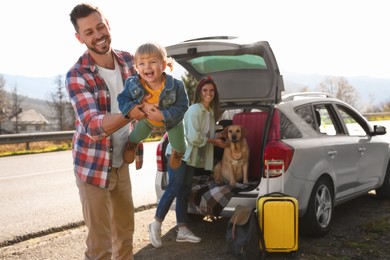 Photo of Father playing with his daughter, happy woman and dog sitting in car trunk near road. Family traveling with pet