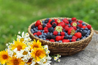 Photo of Wicker bowl with different fresh ripe berries and beautiful flowers on wooden surface outdoors