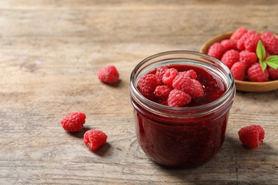 Photo of Glass jar of sweet jam with ripe raspberries on wooden table. Space for text