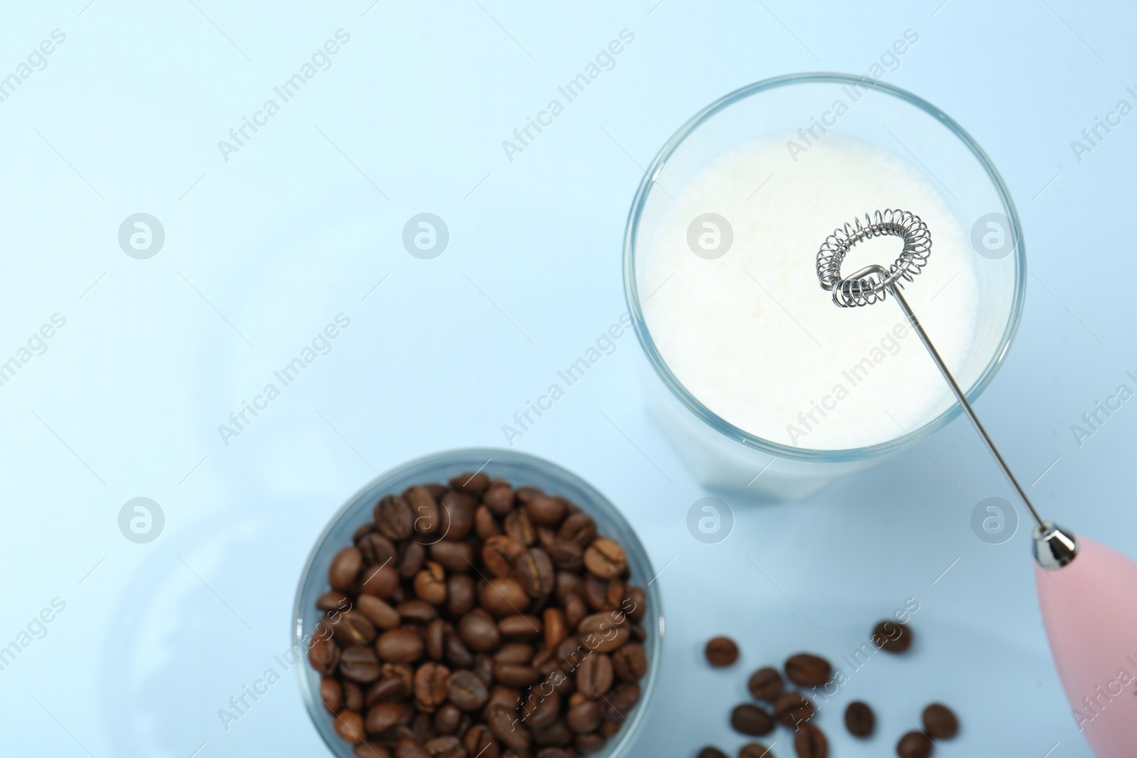 Photo of Mini mixer (milk frother), whipped milk and coffee beans in glasses on light blue background, flat lay. Space for text