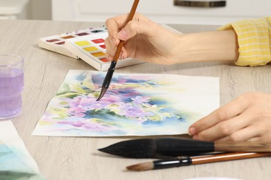 Woman painting flowers with watercolor at white wooden table indoors, closeup. Creative artwork