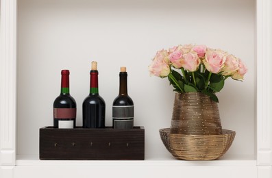 Shelf with different wine bottles and beautiful rose flowers indoors