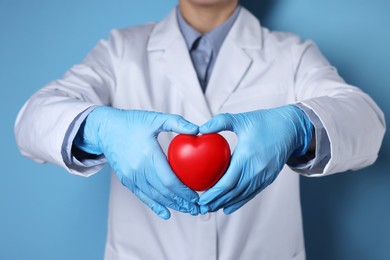 Doctor wearing medical gloves holding decorative heart on light blue background, closeup