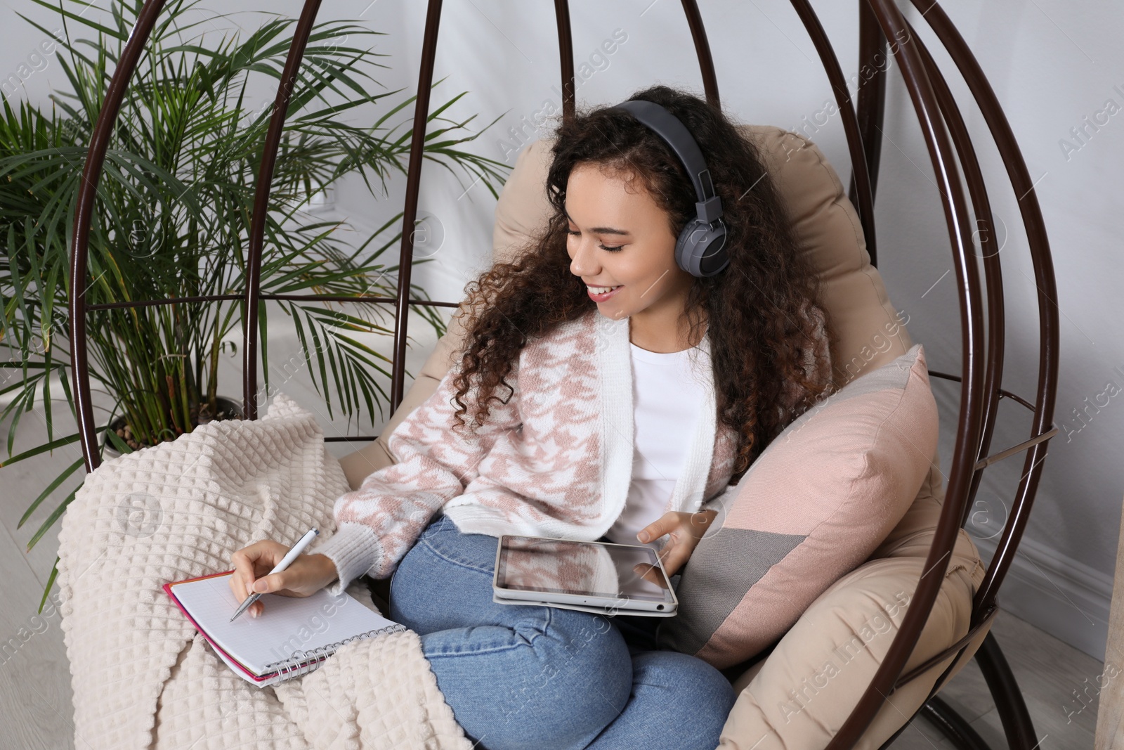 Photo of African American woman with headphones and tablet studying in egg chair at home. Distance learning