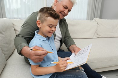 Photo of Little boy with his grandfather solving sudoku puzzle on sofa at home