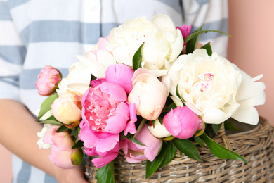 Woman with bouquet of beautiful peonies in basket on beige background, closeup