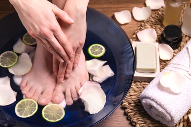 Woman soaking her feet in bowl with water, petals and lime slices on floor, closeup. Spa treatment