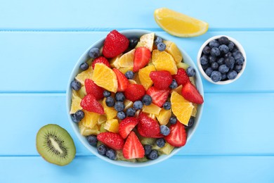 Delicious fresh fruit salad in bowl and ingredients on light blue wooden table, flat lay