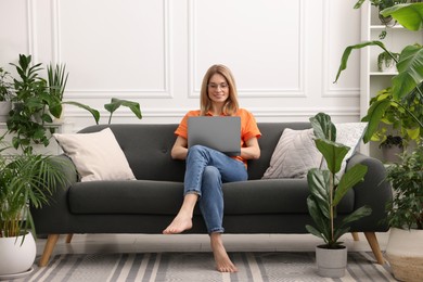 Photo of Woman working with laptop on sofa surrounded by beautiful potted houseplants at home