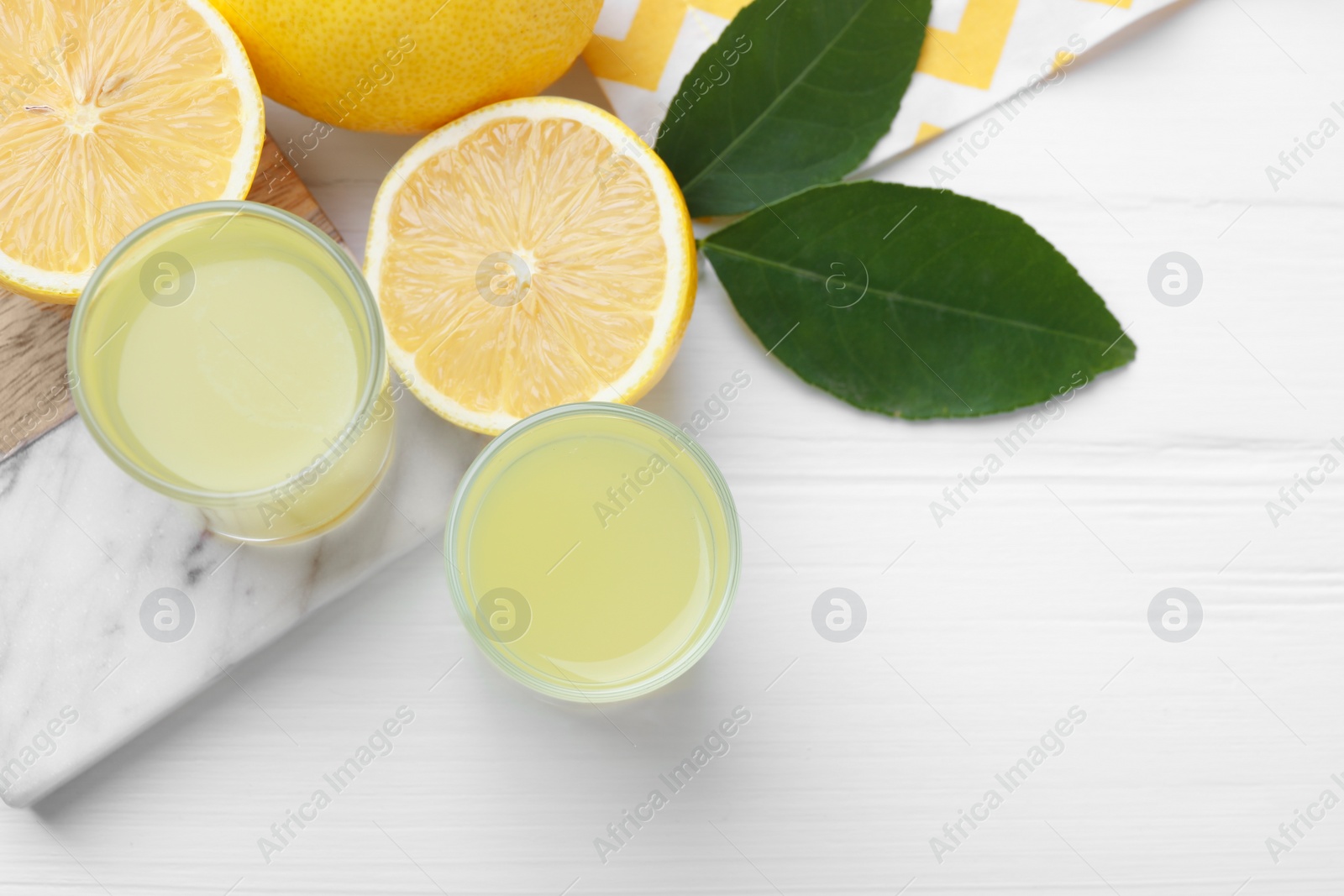 Photo of Tasty limoncello liqueur, lemons and green leaves on white wooden table, top view. Space for text