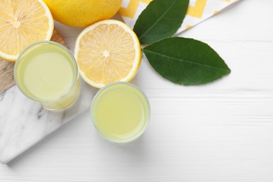Photo of Tasty limoncello liqueur, lemons and green leaves on white wooden table, top view. Space for text