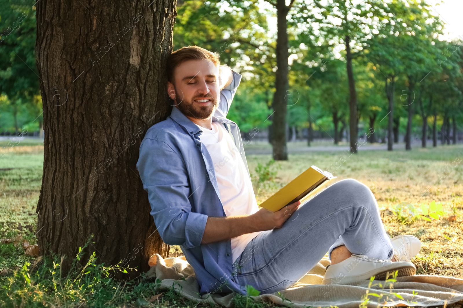 Photo of Young man reading book on green grass near tree in park