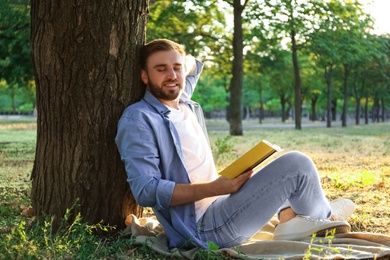 Young man reading book on green grass near tree in park