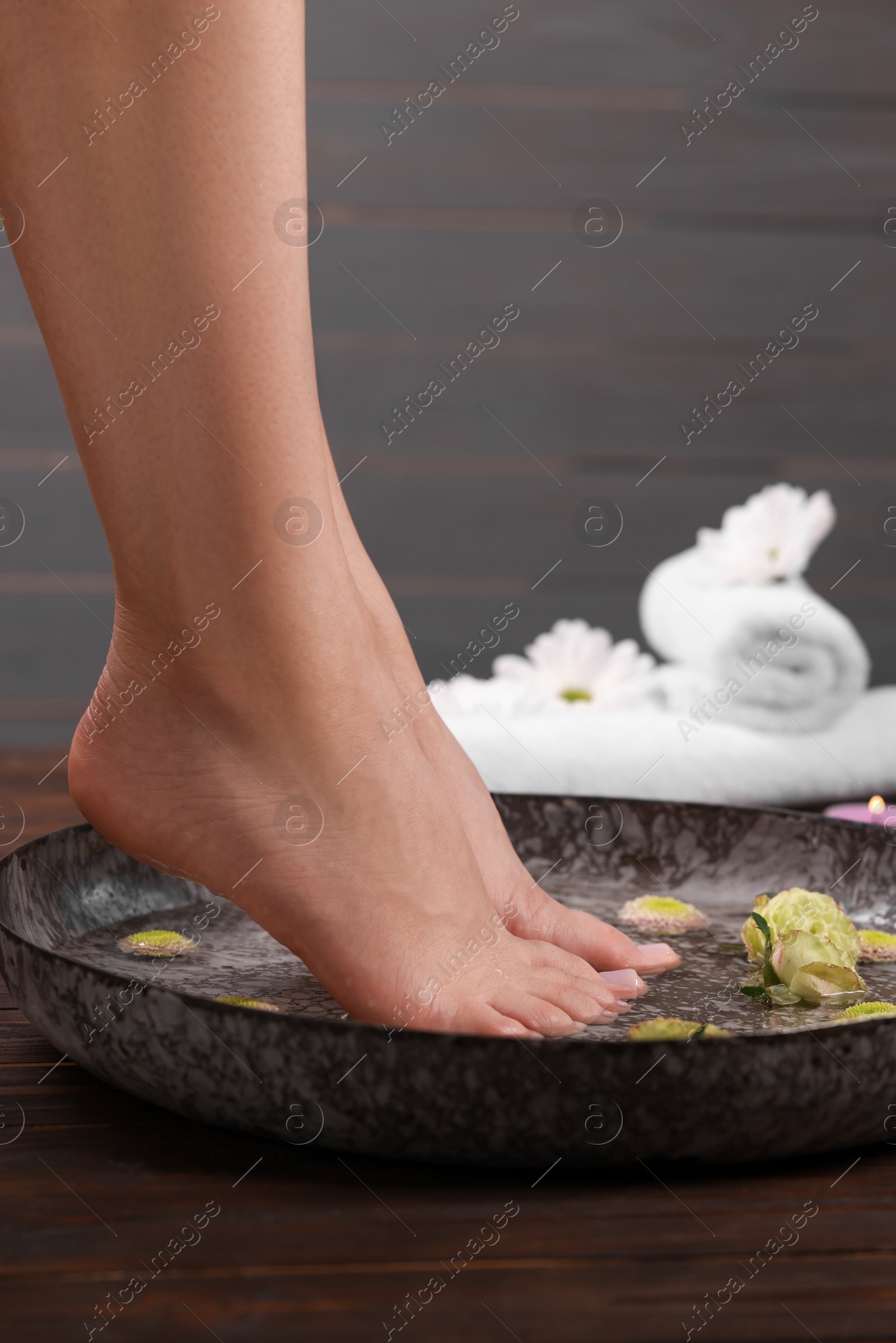 Photo of Woman soaking her feet in bowl with water and flowers on wooden surface, closeup. Pedicure procedure