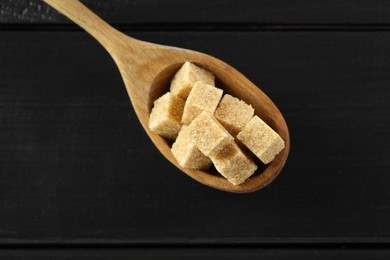 Photo of Brown sugar cubes in spoon on black wooden table, top view