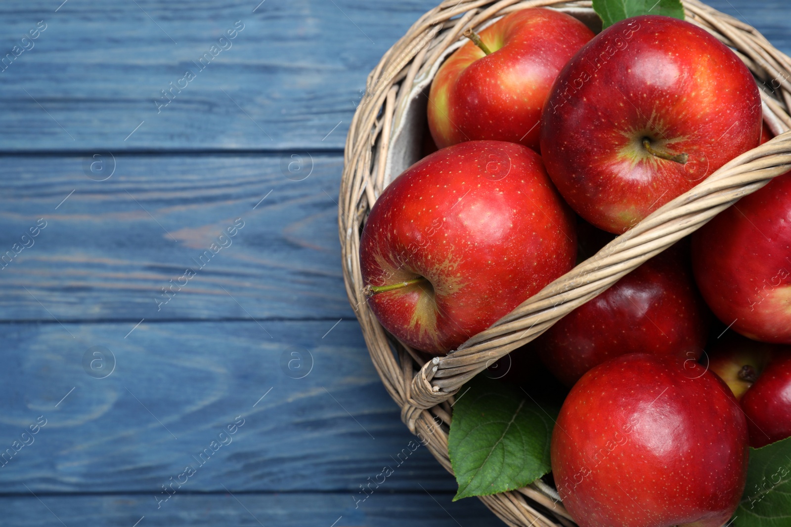Photo of Juicy red apples in wicker basket on blue wooden table, top view