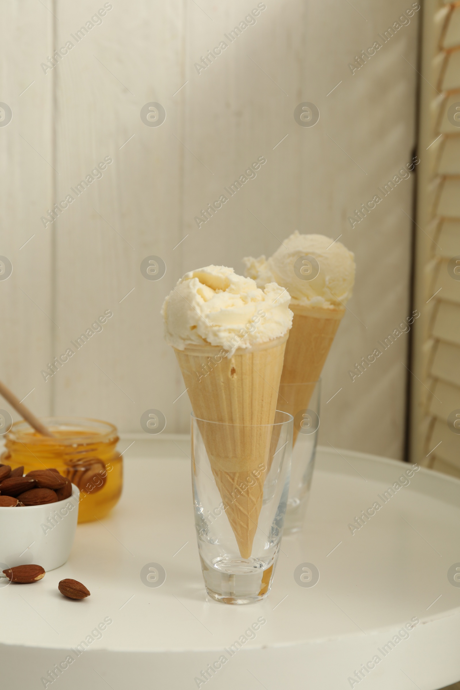 Photo of Delicious vanilla ice cream in wafer cones with honey and almonds on white table indoors