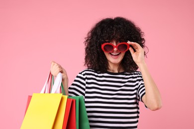 Happy young woman with shopping bags and stylish sunglasses on pink background