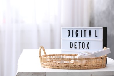 Photo of Gadgets in wicker basket near lightbox with phrase DIGITAL DETOX on white table indoors. Space for text