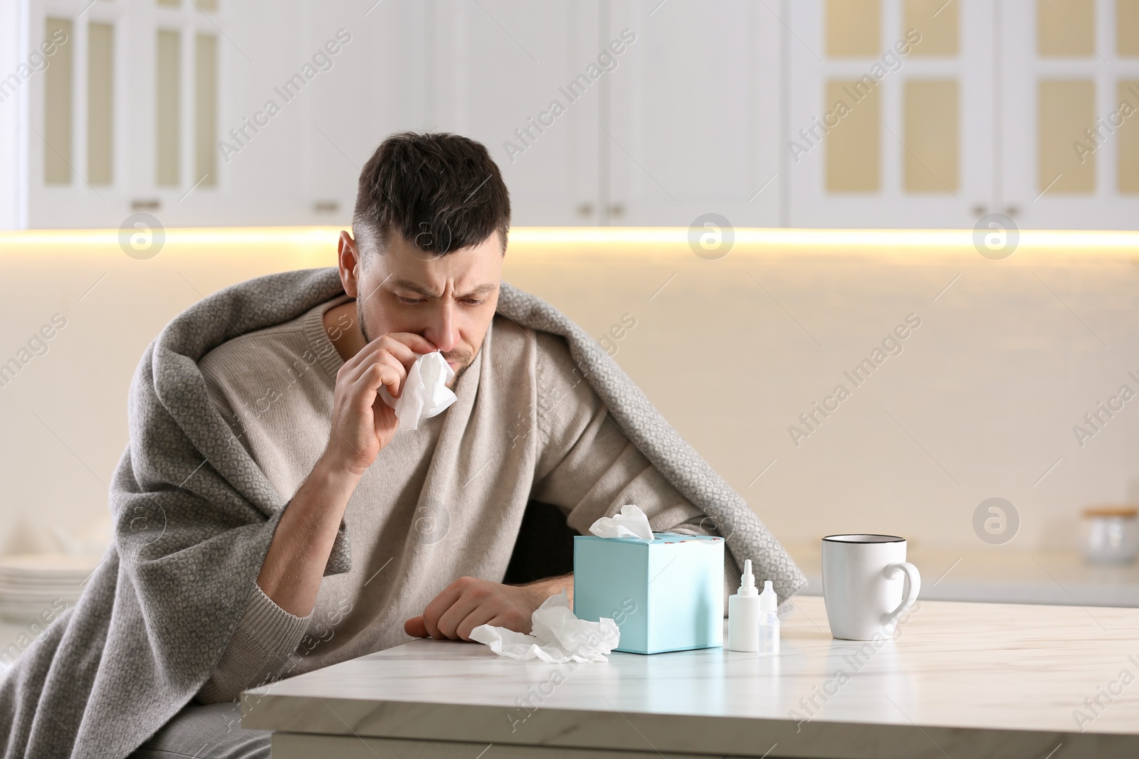 Photo of Ill man at table with nasal spray, drops and box of paper tissues in kitchen