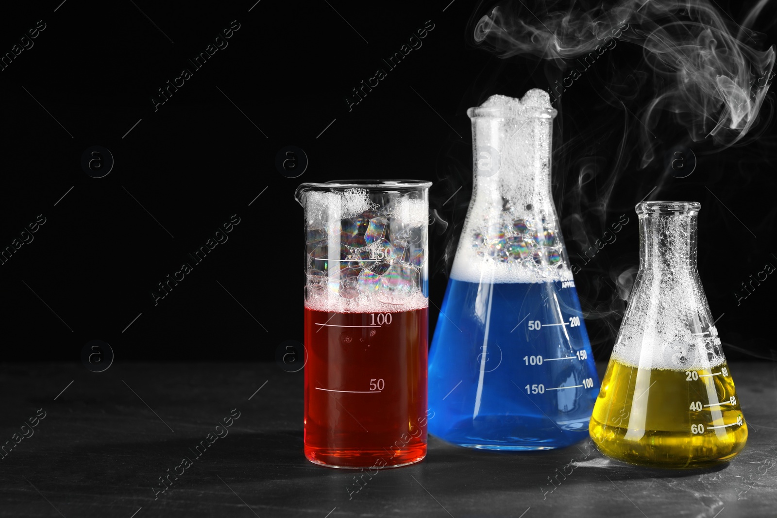 Photo of Laboratory glassware with colorful liquids and steam on dark table against black background, space for text. Chemical reaction